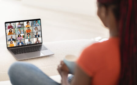 A woman sits in front of an open laptop, participating in a video conference meeting. She is engaged in the meeting and appears to be listening intently to the speakers.